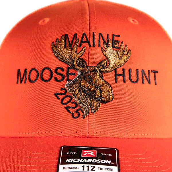 Orange cap with 'Maine Moose Hunt 2025' text and moose graphic on a mannequin head against a white background. Close-up of front of the hat showing how detailed the moose is. 