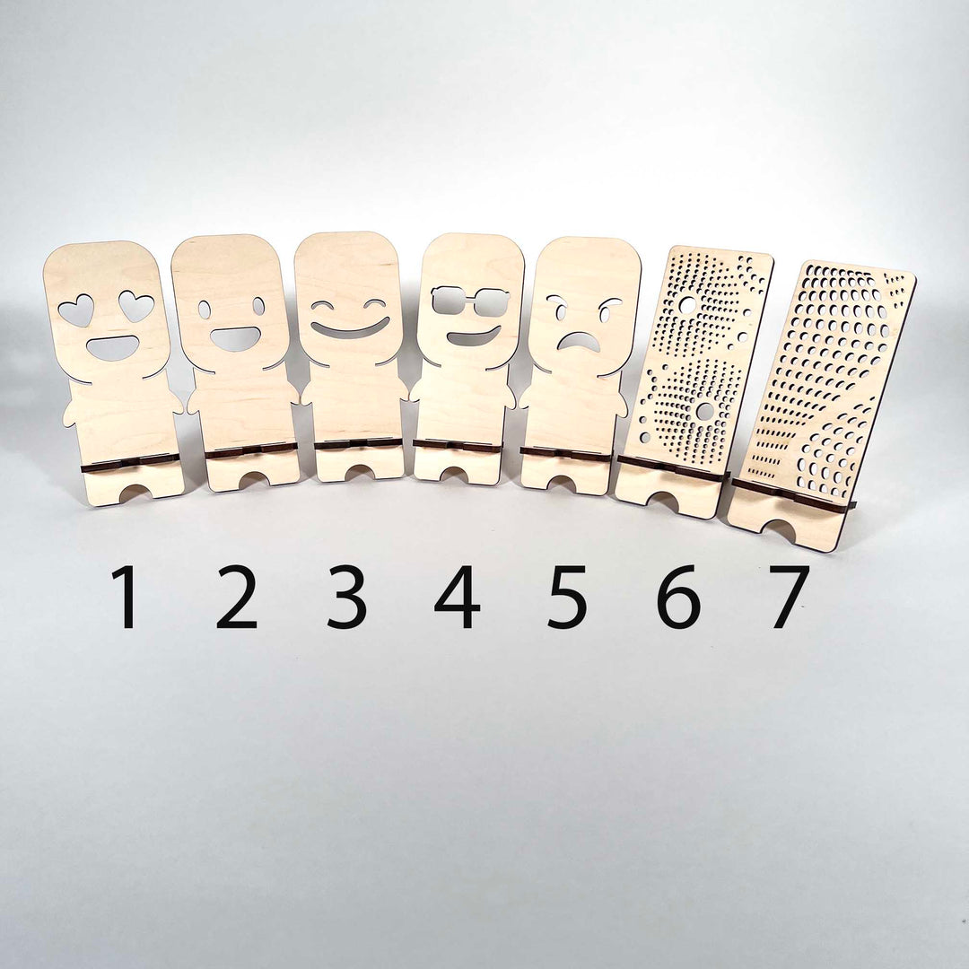 Seven phone stands made from 5mm (approximately 1/4") Birch plywood and the unique designs are laser cut. The designs are #1 Heart Eyes #2 Astonished #3 Smiling #4 Cool Dude #5 Grumpy #6 Starburst #7 Wave - Dailey Woodworking