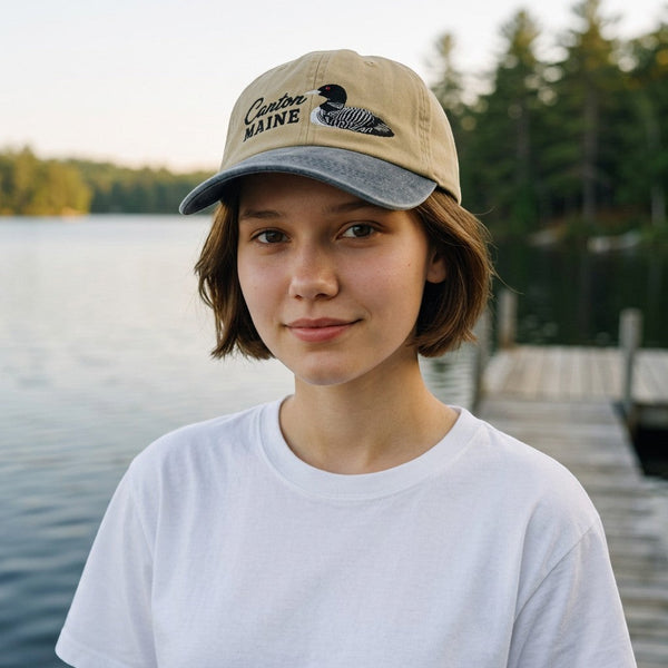 Dailey Woodworking Canton Maine hat with loon embroidery, featuring a tan crown and grey brim on a woman by a lake.