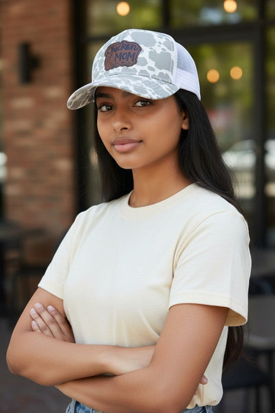 Dailey Woodworking grey cow print trucker hat with white mesh back and "Chicken Mom" leatherette patch. Features ponytail opening.