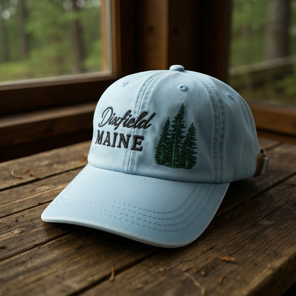Dailey Woodworking light blue cotton hat with Dixfield Maine script and pine tree embroidery on a wooden table.