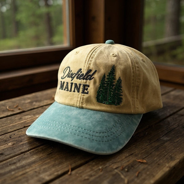 Dailey Woodworking tan and blue garment-washed cotton hat with Dixfield Maine pine tree embroidery on a wooden table.