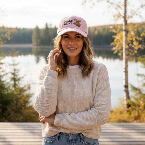 Woman wearing a pink Dailey Woodworking cotton dad hat with Dixfield, Maine and bull moose embroidery by a lake.