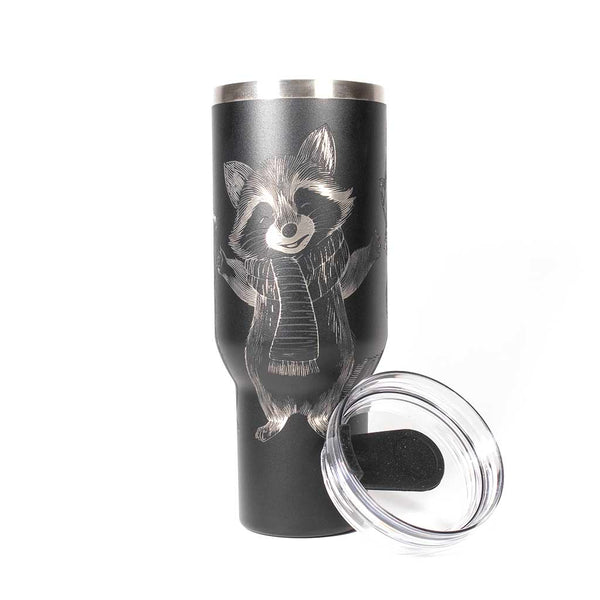 40 ounce black travel mug with three engraved raccoons dressed in scarves and hats. With cover leaned up against tumbler. 