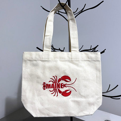 A small canvas gift tote bag embroidered with a red lobster that spells out Maine in the body of the lobster. - Dailey Woodworking